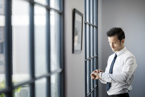 Geschäftsmann überprüft seine Smartwatch vor einem großen Bürofenster, lizenzfreies Stockfoto