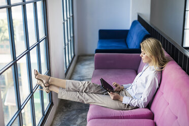 Blond businesswoman sitting on violet couch at the window using tablet - SBOF01582