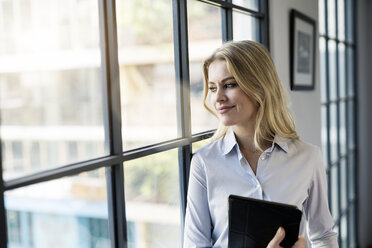 Confident businesswoman with tablet in office looking out of window - SBOF01583