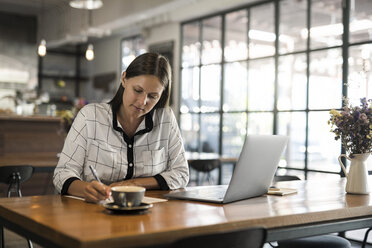 Junge Geschäftsfrau in einem Café schreibt auf Papier und arbeitet mit einem Laptop auf einem Holztisch - SBOF01585