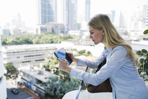 Blonde Frau mit Tablet lehnt sich an ein Glasgeländer in der Stadt, lizenzfreies Stockfoto