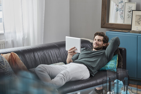 Smiling man with tablet lying on couch at home stock photo