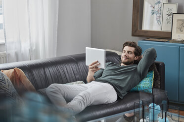 Smiling man with tablet lying on couch at home - RORF01572