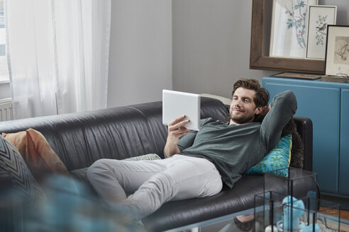 Smiling man with tablet lying on couch at home - RORF01572