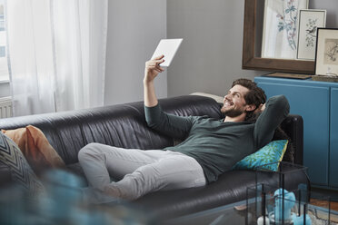 Smiling man with tablet lying on couch at home - RORF01576