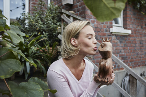 Junge Frau sitzt auf einer Treppe und küsst einen hölzernen Froschprinzen - RORF01648