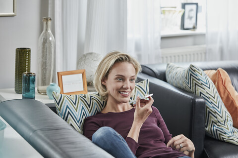 Lächelnde junge Frau, die ein Mobiltelefon benutzt und zu Hause auf der Couch liegt, lizenzfreies Stockfoto