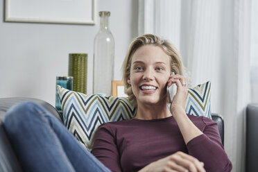 Lächelnde junge Frau mit Handy auf der Couch zu Hause liegend - RORF01658
