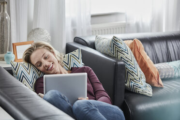 Smiling young woman with tablet lying on couch at home - RORF01659