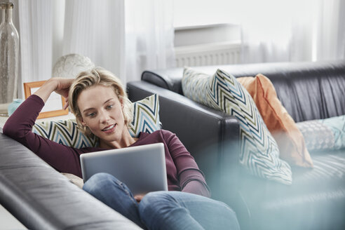 Junge Frau mit Tablet auf der Couch zu Hause liegend - RORF01660