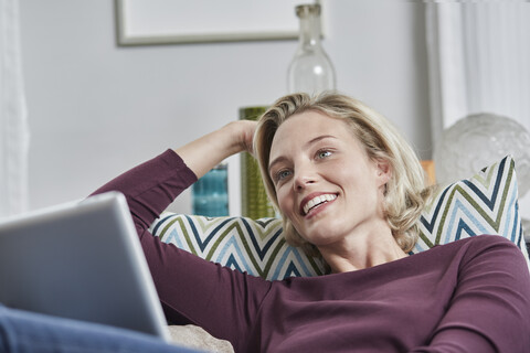 Lächelnde junge Frau mit Tablet auf der Couch zu Hause liegend, lizenzfreies Stockfoto