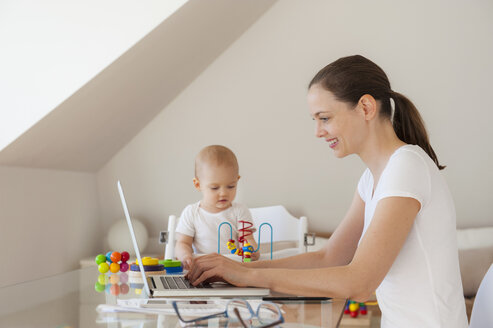 Lächelnde Mutter mit Laptop und kleiner Tochter, die zu Hause am Tisch spielt - DIGF05613