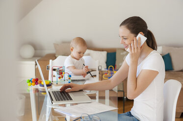 Lächelnde Mutter mit Laptop und Handy mit kleiner Tochter spielen am Tisch zu Hause - DIGF05618
