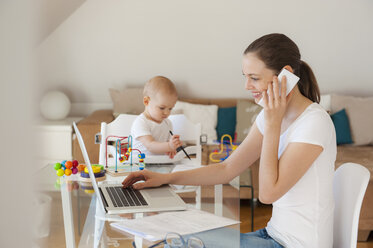 Smiling mother using laptop and cell phone with little daughter playing at table at home - DIGF05618