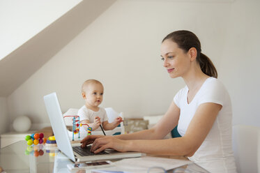 Smiling mother using laptop and little daughter playing at table at home - DIGF05621