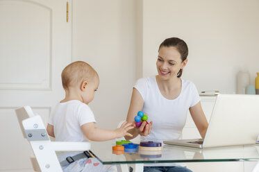 Smiling mother using laptop playing with little daughter at table at home - DIGF05622