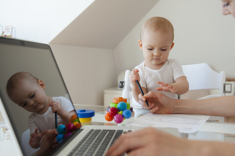 Kleine Tochter spielt und Mutter benutzt Laptop am Tisch zu Hause, lizenzfreies Stockfoto