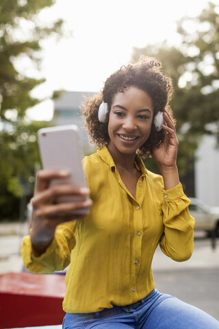 Porträt einer lächelnden Frau, die mit Kopfhörern Musik hört und ein Selfie mit ihrem Smartphone macht, lizenzfreies Stockfoto