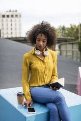 Woman sitting on bench with coffee to go and notebook looking at smartphone - MAUF02334