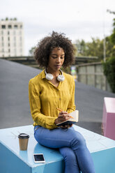 Woman sitting on bench with smartphone and coffee to go writing in notebook - MAUF02335