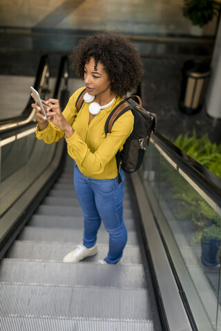 Frau mit Rucksack und Kopfhörern steht auf der Rolltreppe und benutzt ihr Smartphone, lizenzfreies Stockfoto