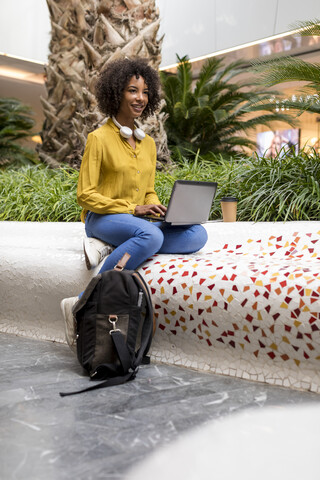 Porträt einer lächelnden Frau, die auf einer Bank sitzt und einen Laptop benutzt, lizenzfreies Stockfoto