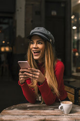 Porträt einer lachenden jungen Frau, die auf der Terrasse eines Cafés sitzt und ihr Smartphone benutzt - ACPF00372