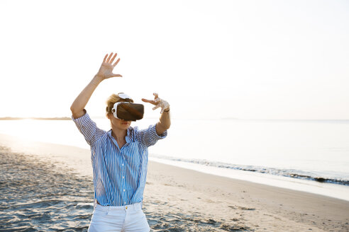 Blonde woman doing kind of yoga exercises on a beach in thailand with 3D virtual reality goggles - HMEF00177
