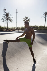 Muskulöser Mann mit nacktem Oberkörper bei Dehnungsübungen in einem Skatepark - MAUF02373