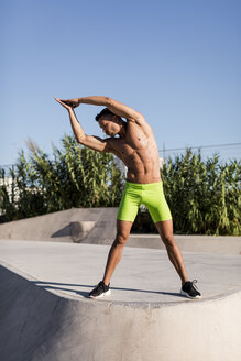 Muskulöser Mann mit nacktem Oberkörper bei Dehnungsübungen in einem Skatepark - MAUF02382