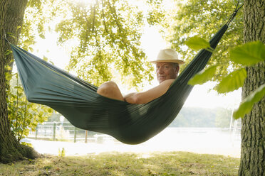 Senior man wearing straw hat relaxing in hammock at lakeshore - GUSF01783