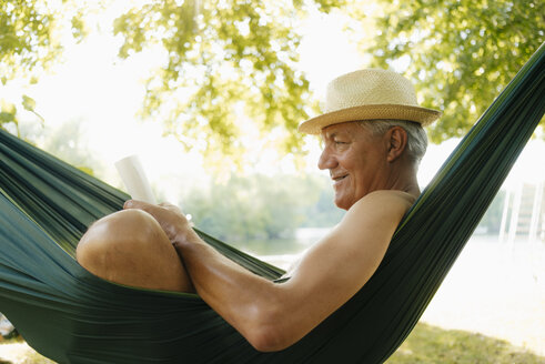 Älterer Mann mit Strohhut entspannt sich in einer Hängematte am Seeufer und liest ein Buch - GUSF01788