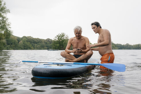 Älterer und jüngerer Mann mit SUP-Board auf einem See und Mobiltelefon - GUSF01802