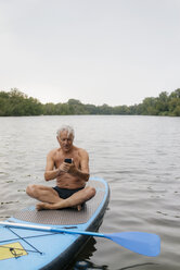 Älterer Mann sitzt auf einem SUP-Board auf einem See und benutzt ein Mobiltelefon - GUSF01803