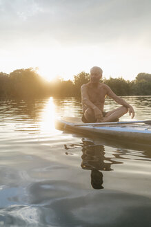 Älterer Mann sitzt bei Sonnenuntergang auf einem SUP-Board - GUSF01816