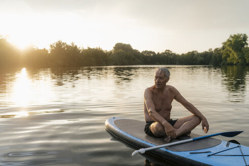 Älterer Mann sitzt bei Sonnenuntergang auf einem SUP-Board - GUSF01817