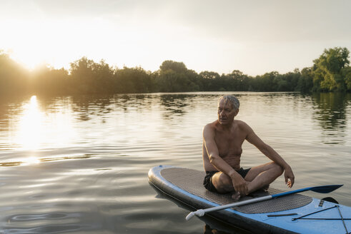 Senior man sitting on SUP board at sunset - GUSF01817