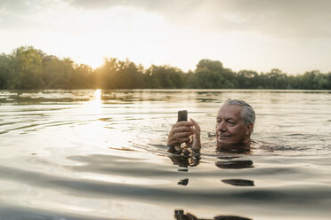 Senior man swimming in a lake at sunset using cell phone - GUSF01823