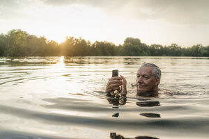 Älterer Mann, der bei Sonnenuntergang in einem See schwimmt und ein Handy benutzt - GUSF01823