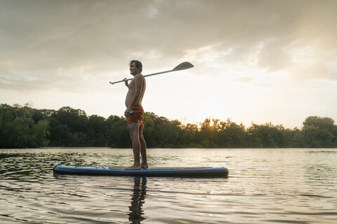 Mann steht auf SUP-Board auf einem See bei Sonnenuntergang, lizenzfreies Stockfoto