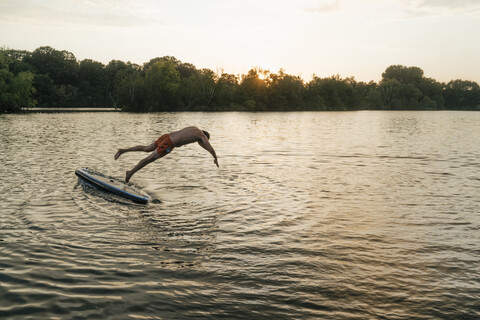 Mann springt von SUP-Board auf einem See bei Sonnenuntergang, lizenzfreies Stockfoto