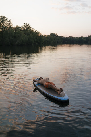 Mann liegend auf SUP-Board auf einem See bei Sonnenuntergang, lizenzfreies Stockfoto