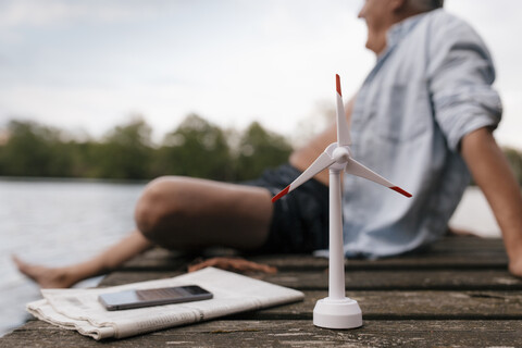 Älterer Mann sitzt auf einem Steg an einem See mit einem kleinen Windradmodell, lizenzfreies Stockfoto