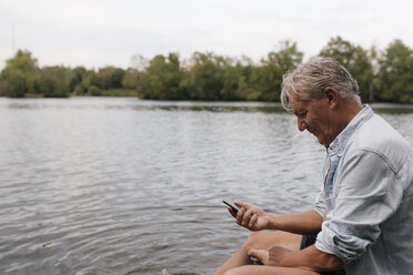 Älterer Mann sitzt an einem See und benutzt ein Mobiltelefon - GUSF01844