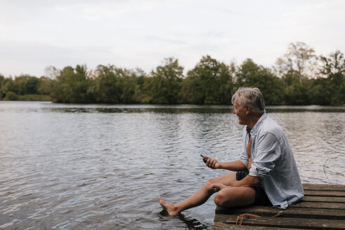 Glücklicher älterer Mann sitzt auf einem Steg an einem See mit Handy - GUSF01845