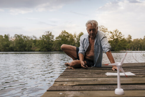 Älterer Mann sitzt auf einem Steg an einem See mit einem kleinen Windradmodell - GUSF01846