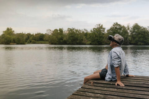 Älterer Mann sitzt auf einem Steg an einem See und trägt eine VR-Brille, lizenzfreies Stockfoto