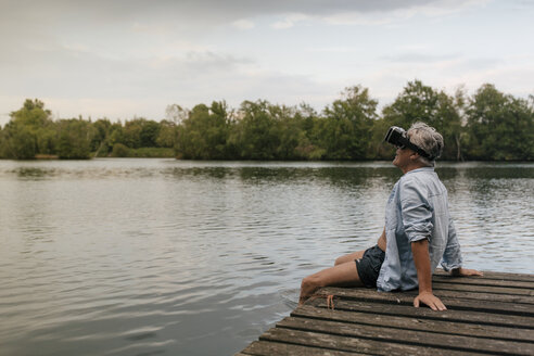 Älterer Mann sitzt auf einem Steg an einem See und trägt eine VR-Brille - GUSF01847
