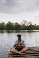 Älterer Mann sitzt auf einem Steg an einem See, trägt eine VR-Brille und trinkt Kaffee - GUSF01849