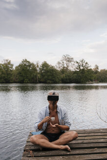 Älterer Mann sitzt auf einem Steg an einem See, trägt eine VR-Brille und trinkt Kaffee - GUSF01849